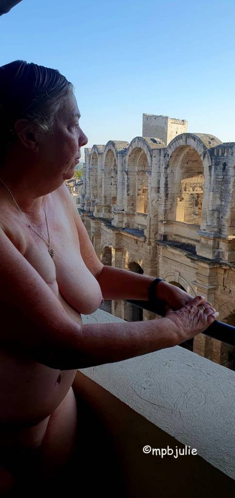 I am standing naked on a balcony, hands resting on the railing. I am sideways on and you can see my breasts, arms and tummy. Across the street is the remains of an ancient arena. 