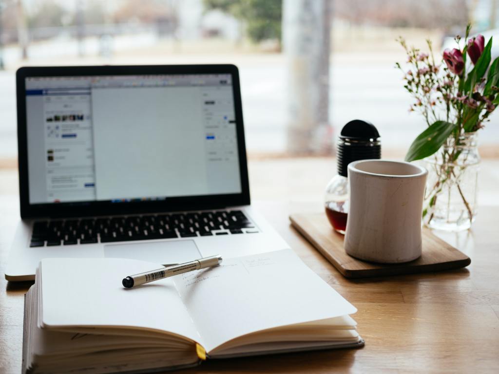 A desk containing laptop, book and pen and coffee.