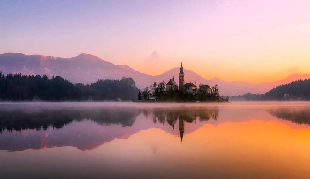 body of water near mountain during daytime with a church at the centre. The image is reflected into the water 