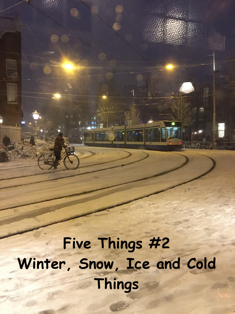 A winter snowy scene in Amsterdam. A cyclist is waiting to cross the tracks as a tram approaches. It is night. The now on the ground shines brightly.