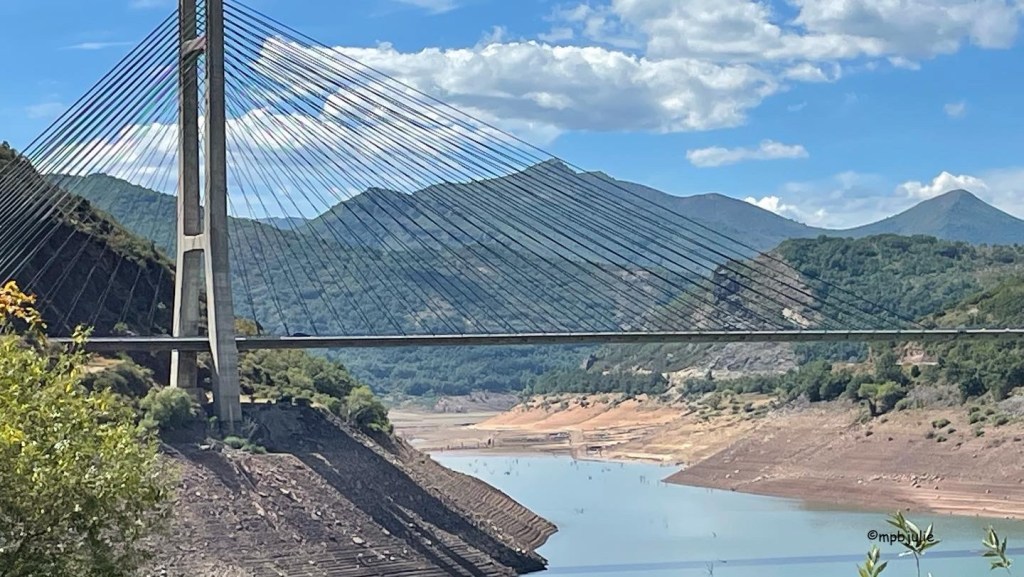 There is a suspension bridge in the foreground over a lake with a sandy river shore. There are mountains and. hills in the background, the lower ones covered in greenery. The sky is blue with a few clouds