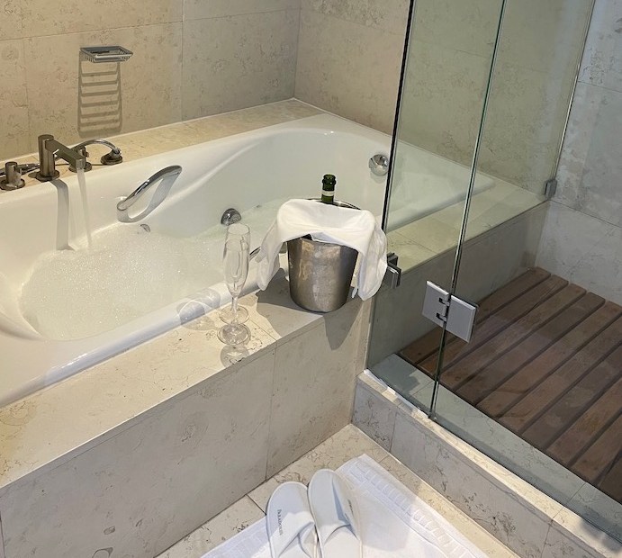 The image shows a bath filling with water and a few bubbles forming. On the side of the bath is a wine bucker containing a bottle of cava, covered with a towel. There are two glasses (plastic). On the floor is a white mat and some parador slippers. To the right you can see there is a separate shower in this bathroom.