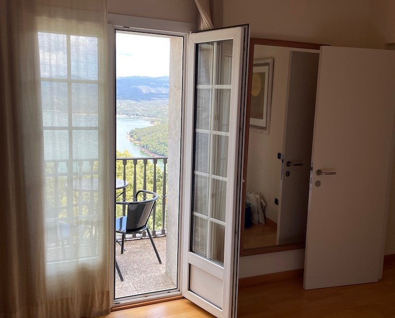 Room with doors, one open. Outside is a chair and railings then a lake and trees.