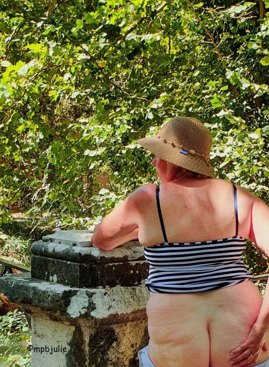 I'm standing leaning on a weathered stone post with greenery from trees in the background. I'm wearing a sun hat and blue and white stripy sun top, pulled up to my waist. My bottom is bare and my hand rests on my right butt cheek. I'm facing the bushes.