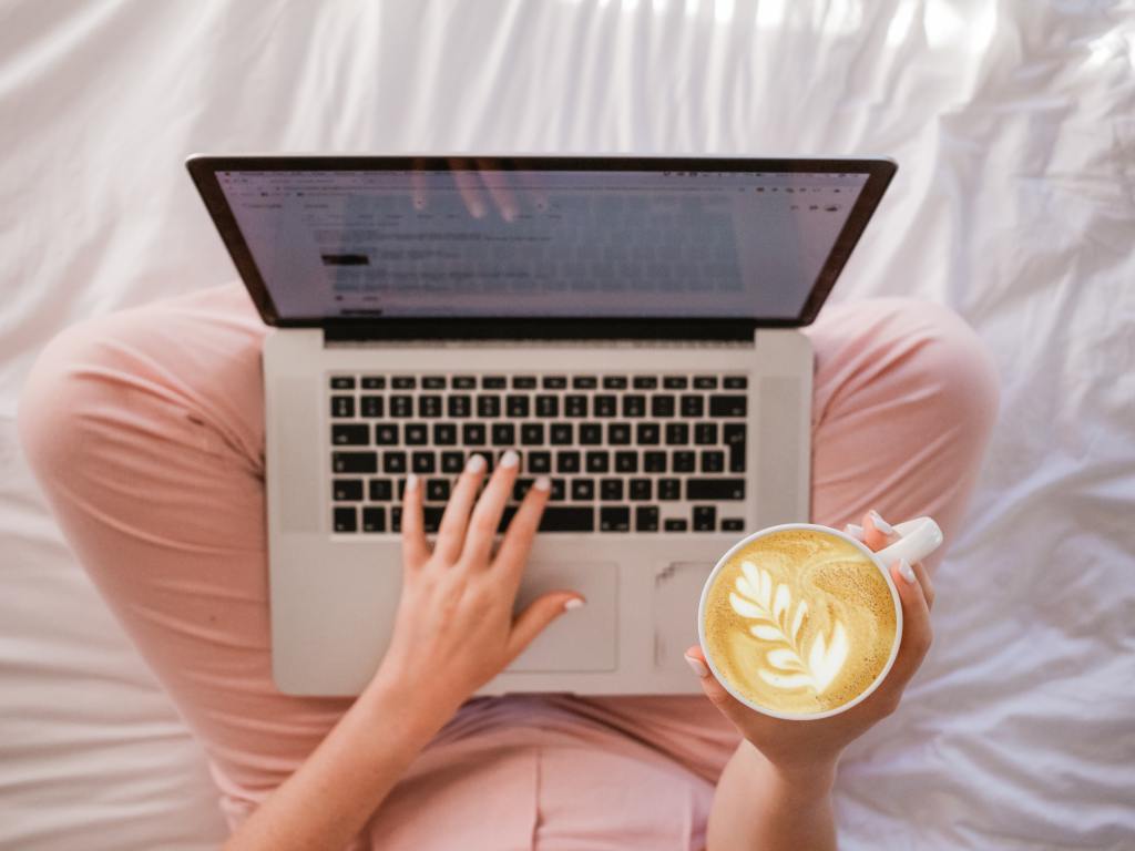 Someone sitting cross legged on a bed with laptop and coffee. 