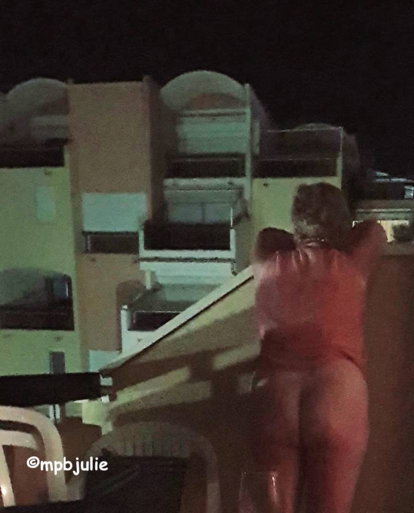 Photo of me standing wearing just my top facing away from the camera. I'm leaning on the side balcony and looking at the apartments opposite.