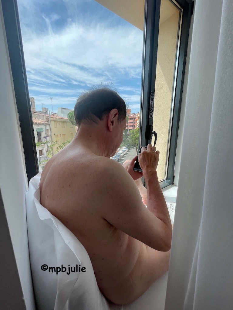 Master is sitting naked on a huge window sill. He is behind the curtains and leaning against a pillow looking at his phone.
