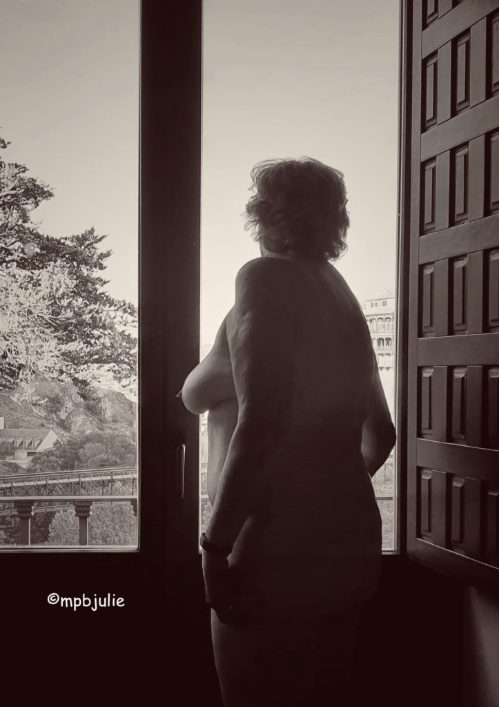 I'm standing in front of a large window with a wooden shutter to the right. In the foreground is a large tree, hills and a building and bridge. The image has been altered so it is black and white. you can see my side view or back and bottom and left breast. I'm naked.