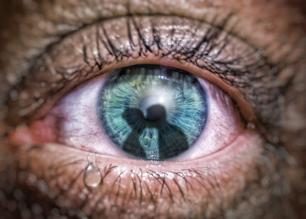 An eye with a tear on the bottom lid. A couple kissing are reflected in the iris. 
