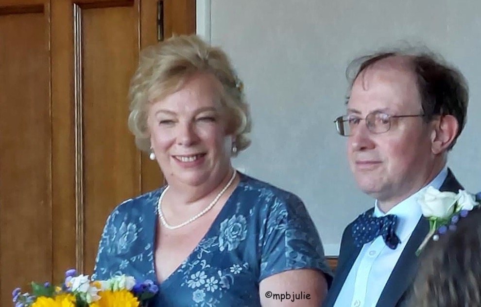 A photo of us both as we are announced as man and wife. I am wearing a blue dress with pale blue blowers.  I am carrying a bouquet containing yellow, white and blue flowers. Master is wearing a blue bow tie and white shirt under his black jacket. he is wearing a white rose.